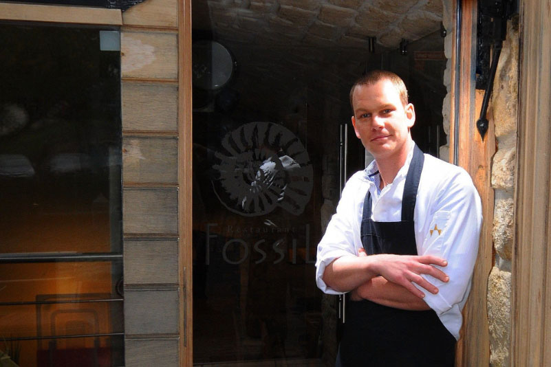 Junger Koch vor Restaurant-Eingang mit Logo, trägt Kochjacke und schwarze Schürze.