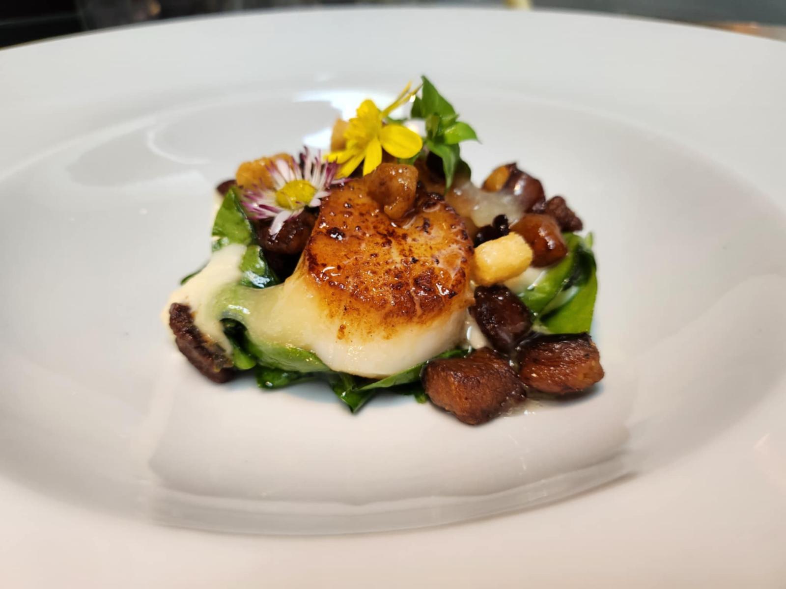 Gebratene Jakobsmuschel mit Gemüse, Käse, Nüssen und essbaren Blumen auf weißem Teller.